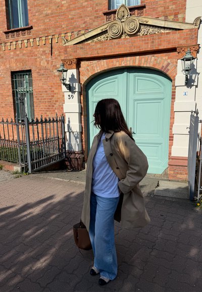 Mujer con cabello largo y oscuro que lleva un abrigo beige, camisa blanca y vaqueros azules, de pie en la acera cerca de unas puertas dobles de color verde claro de un edificio de ladrillo rojo.