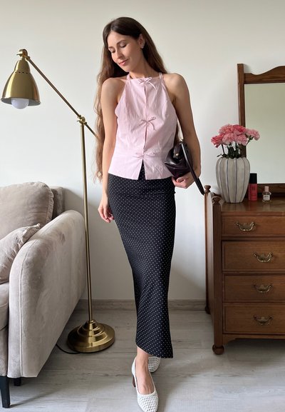 Top halter rosa claro con lazos, combinado con una falda negra de lunares. La modelo sostiene un bolso oscuro, de pie junto a una lámpara decorativa y una cómoda.