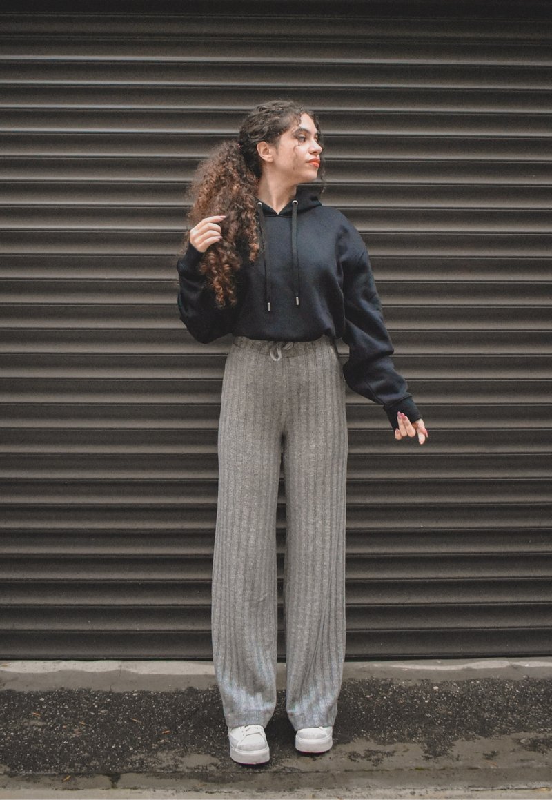 Black hooded sweatshirt with drawstrings, paired with grey ribbed wide-leg trousers. White sneakers. Background features a textured dark surface.