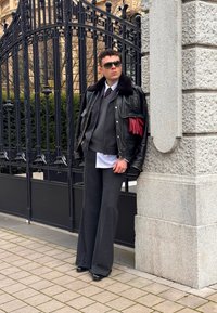 Homme portant des lunettes de soleil, une veste en cuir avec des gants rouges, un pull gris et un pantalon à jambes larges, appuyé contre un mur de pierre près d'une clôture en fer noir.