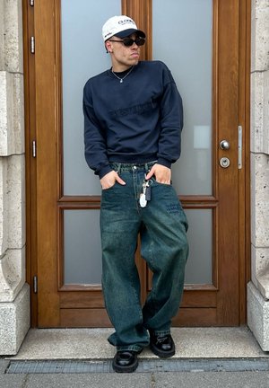 Sudadera marina con letras texturizadas, pantalones de denim verdes de gran tamaño, zapatos negros gruesos, gorra blanca con logo y gafas de sol, posado junto a una puerta.