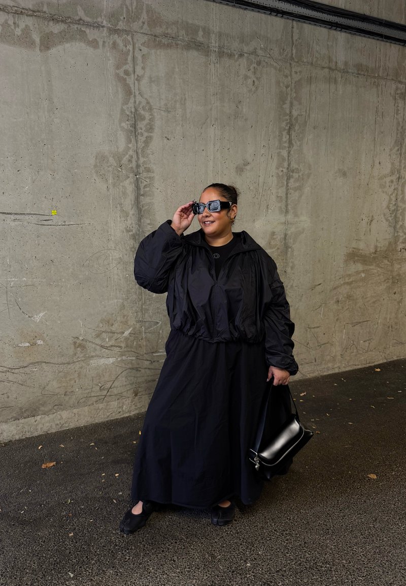 Black oversized jacket with gathered waist, black long dress, and black flat shoes. Accessories include dark sunglasses and a small black bag.