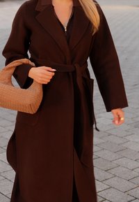 Καφέ μάλλινο παλτό με ζώνη στη μέση, μεγάλα πέτα και πλαϊνές τσέπες. Κρατά μια πλεκτή καφέ τσάντα. Εξωτερικός χώρος με πέτρινο πλακόστρωτο.