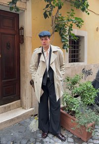 Bež trench coat preko bijele košulje, tamno prugastih hlača i bordo cipela. Sivi šešir upotpunjuje izgled, dok u pozadini ima zelenila.