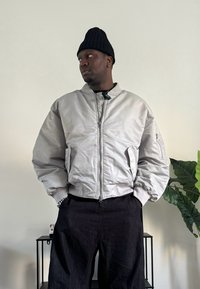 Light gray bomber jacket with a zip front, ribbed cuffs, and a pocket on the sleeve, paired with black pants and a black beanie.