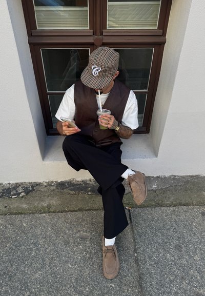 Chaleco de cuero marrón sobre una camiseta blanca, pantalones anchos negros, zapatos beige, sosteniendo una bebida y un teléfono, sentado junto a una ventana.