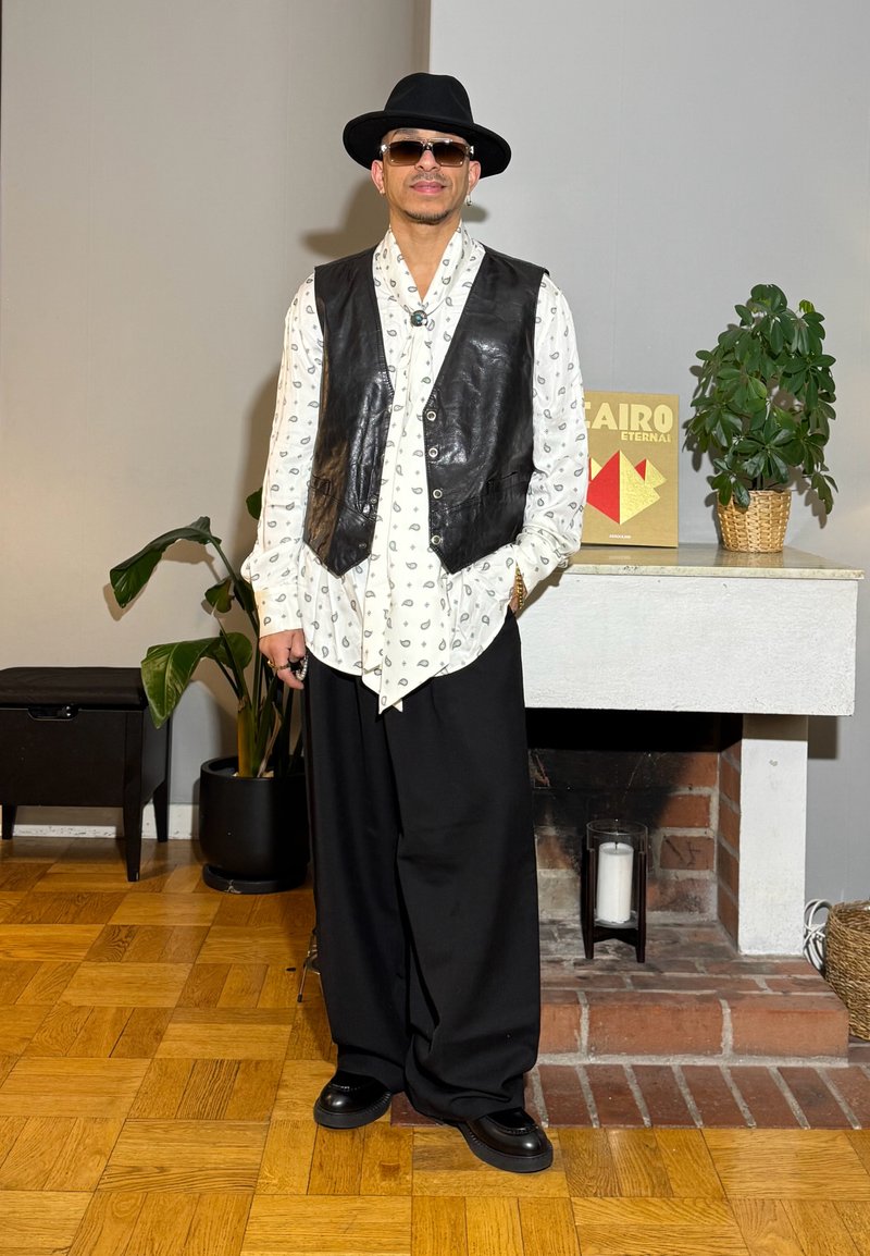 Homme portant un chapeau noir, des lunettes de soleil, une chemise blanche à motifs, un gilet en cuir noir et un pantalon large noir, debout à l'intérieur près de plantes et d'une cheminée.