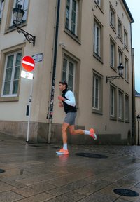 Un coureur portant un débardeur noir et un short gris, chaussé de baskets orange et blanches, fait son jogging sur une rue pavée mouillée.