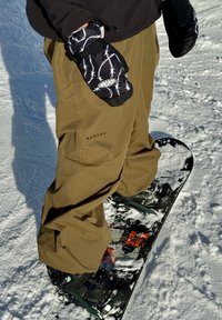 Personne debout sur un snowboard couvert de neige portant un pantalon Oakley marron et des moufles noires avec un motif de fil barbelé blanc.