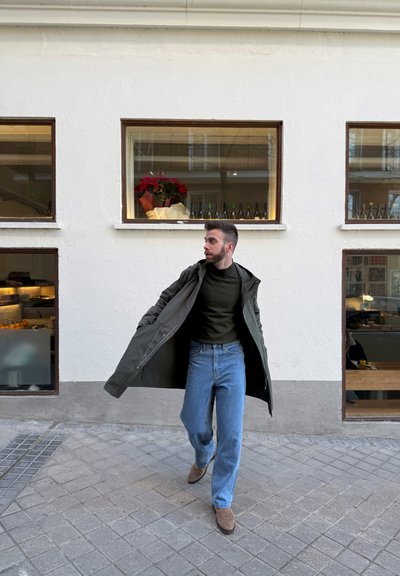 Abrigo largo verde, suéter texturizado verde oscuro, jeans de pierna ancha azules y botas de ante marrones. Entorno urbano con pared blanca y grandes ventanales.