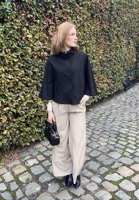 Top corto nero con colletto alto; pantaloni beige larghi; borsa nera; pavimentazione in pietra testurizzata; sfondo verdastro di fogliame.