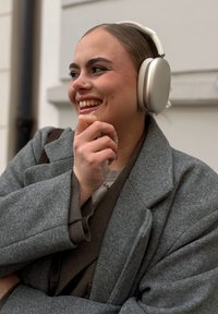 Draadloze over-ear koptelefoon met een zilveren afwerking en zachte witte padding, gedragen door een persoon in een grijze jas, die glimlacht en een hand op de kin rust.