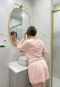 Top rosa a manica lunga e pantaloncini abbinati, con finitura testurizzata. Lavabo a ciotola in ceramica bianca contro una parete a motivo marmo e specchio con accenti dorati.