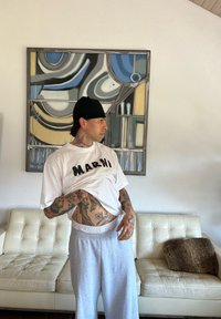 Λευκό μπλουζάκι με μαύρο κείμενο "MARNI", γκρι φόρμες και τατουάζ. Το φόντο περιλαμβάνει έναν αφηρημένο πίνακα και έναν καναπέ σε κρεμ απόχρωση.