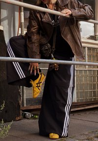 Fekete szalagú sportnadrág, barna bőrdzseki, sárga sneakers és texturált kiegészítők. Az öltözet rétegezést és merész színeket tartalmaz.