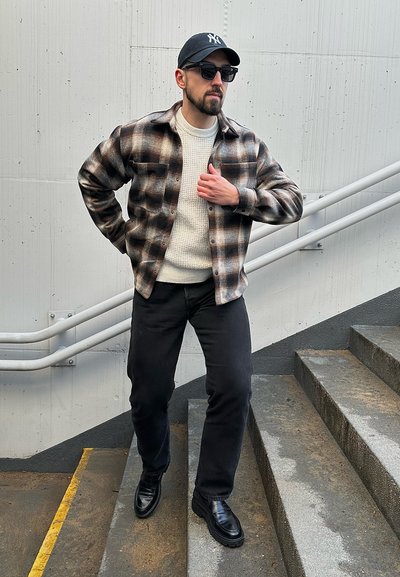 Camisa de franela a cuadros negros y marrones sobre un suéter texturizado crema, combinada con jeans negros y botas negras brillantes, completando un look en capas.