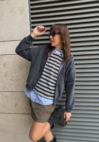Sudadera con capucha gris oscuro con cremallera, usada sobre una camisa a rayas marrón y blanca, combinada con una camisa de cuello azul claro, junto con pantalones cortos oliva y botas negras.