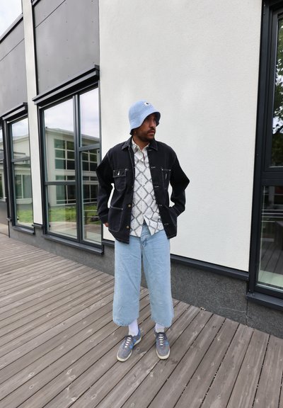 Chaqueta de mezclilla negra con dos bolsillos frontales, camisa de botones estampada, sombrero tipo bucket de color azul claro, vaqueros anchos de color azul claro y zapatillas grises.