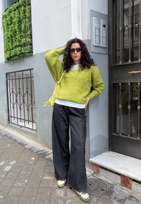 Femme aux cheveux bouclés portant des lunettes de soleil noires, un pull vert citron, un pantalon large noir et des baskets à motifs, debout sur un trottoir près d'un bâtiment.