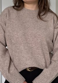 Femme portant un pull en tricot beige avec des poignets côtelés et un col rond, associé à un pantalon noir et une ceinture marron avec une boucle dorée.