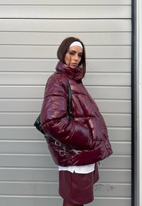 Femme portant une doudoune brillante bordeaux et une jupe avec un bandeau blanc, debout de profil devant un fond de panneaux horizontaux gris.