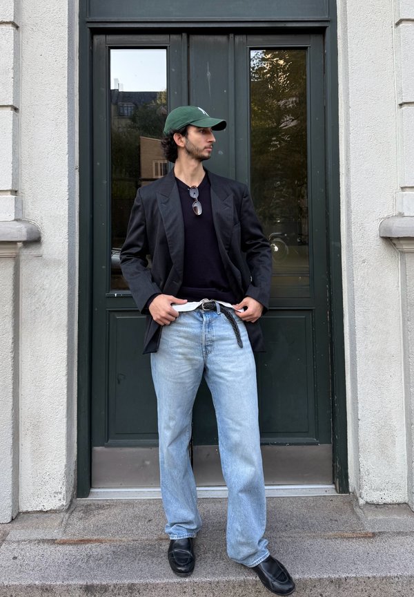 Un hombre con una gorra verde, una chaqueta oscura y unos vaqueros holgados está de pie delante de una puerta verde oscura y mira hacia un lado.