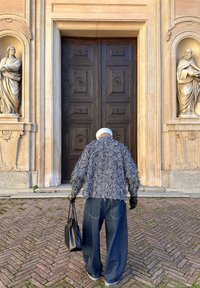 Personne aux cheveux blancs portant une veste texturée noire et blanche et un jean large, tenant un sac noir, debout devant une grande porte en bois foncé.