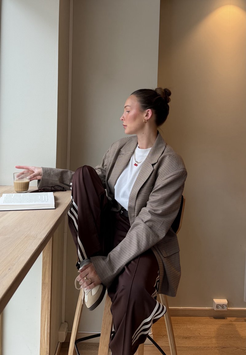 Mujer joven con el cabello recogido en un moño, vestida con un blazer a cuadros y pantalones a rayas, sentada en un taburete junto a la ventana, sosteniendo un vaso y apoyando el pie en una silla.