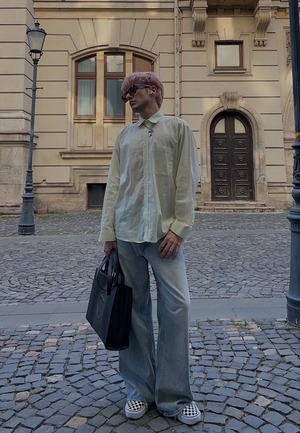Giovane con capelli rosa, occhiali da sole, camicia beige, jeans larghi e scarpe a quadri, in piedi su una strada lastricata con una borsa nera in mano.
