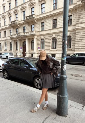 Joven con chaqueta marrón, falda plisada y gorra, de pie junto a un farol en la acera de la ciudad, con coches aparcados y un edificio histórico detrás.