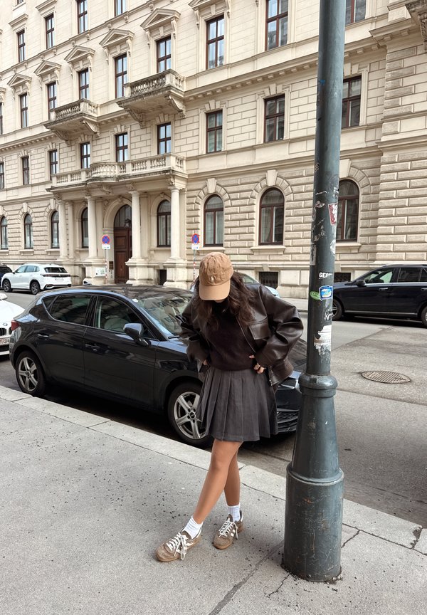 Young woman in brown jacket, pleated skirt, and cap standing near a lamppost on a city pavement with parked cars and a historic building behind.