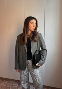 Blazer verde-oliva sobre uma blusa preta, combinado com calças de padrão leopardo em tons claros. Uma pequena bolsa preta com padrão de croco é segurada.