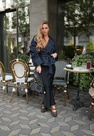 Chaqueta de mezclilla con cintura ajustada con cinturón, combinada con una falda negra ajustada. Bolso negro y zapatos negros con tiras. Mesas redondas y sillas con estampado en el fondo.