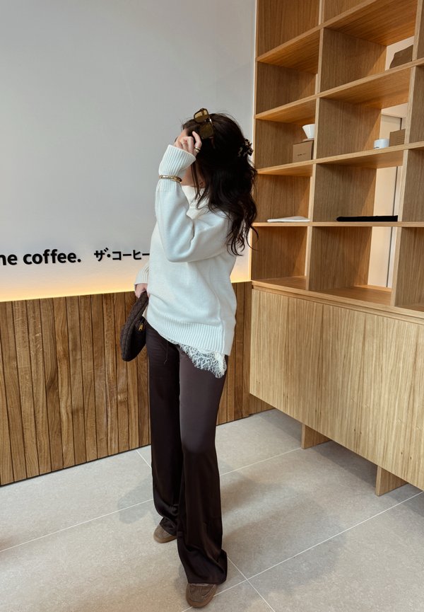 Žena v bielom svetri a hnedých nohaviciach drží kabelku, nastavuje si slnečné okuliare, stojac pri drevených policiach a stenách s obložením a textom.