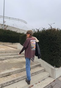 Smeđa grafička majica s tiskanim dizajnom, slojevita preko karo košulje, uparena s trapericama i smeđim čizmama. Bež kapa i crna jakna.