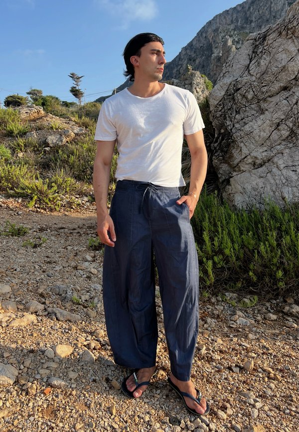 Homme en t-shirt blanc, pantalon large bleu marine et tongs, debout sur un sol rocheux près d’arbustes et de gros rochers sous un ciel dégagé.