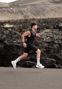 Hardloper in een zwarte mouwloze top en shorts, met witte sokken en sneakers, op een rotsachtig terrein met een geplaveid pad en bergen op de achtergrond.