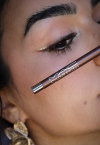 Matita MAC Colour Excess di colore marrone metallico, con punta argentata e logo del marchio. Ingrandimento di un occhio con un accento dorato vicino alla linea delle ciglia.