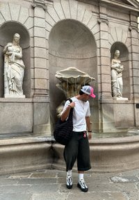 Bolsa acolchada negra, camiseta blanca, pantalones cortos negros hasta la rodilla y zapatillas en blanco y negro, todo ello que se lleva cerca de una fuente de piedra con esculturas clásicas.