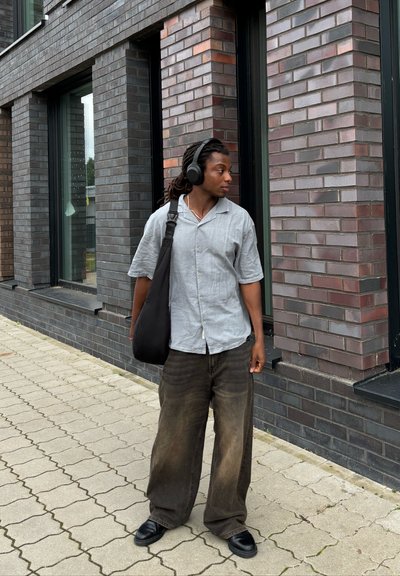 Camisa de botones de manga corta gris, pantalones sueltos marrones, bolso negro y auriculares, de pie contra un edificio de ladrillo en un paseo pavimentado.