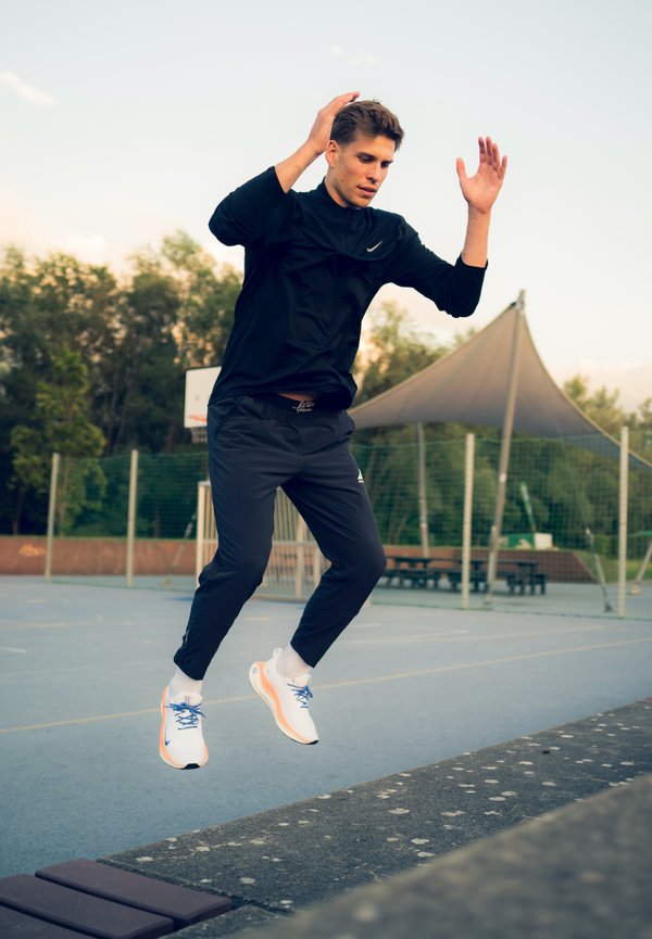 Zapatillas deportivas con un diseño en blanco y naranja, cordones azules; camiseta negra de manga larga y pantalones oscuros; acción de saltar en una cancha deportiva.