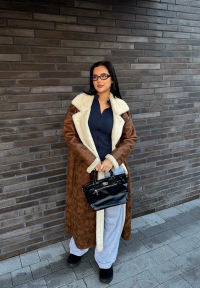 Larga abrigo de piel sintética marrón con forro crema, llevado sobre una camiseta azul marino y pantalones de chándal grises. Un bolso negro y calzado negro grueso completan el conjunto.