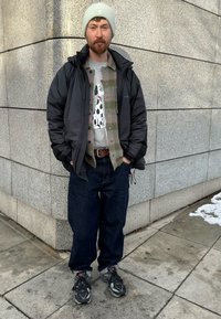 Homme portant une doudoune noire sur un sweat-shirt gris avec un chien de dessin animé, une écharpe à carreaux, un jean foncé et des baskets grises. Neige sur le trottoir.