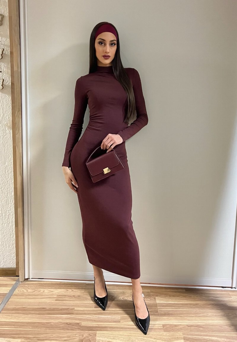 Femme portant une robe longue ajustée à manches longues de couleur bordeaux foncé et un bandeau assorti, tenant un petit sac à main bordeaux, debout sur un sol en bois.