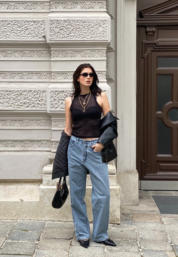 Top crop preto canelado, jeans de perna larga azul claro, jaqueta de couro preta e sapatos de bico fino, complementados com colares dourados em camadas.