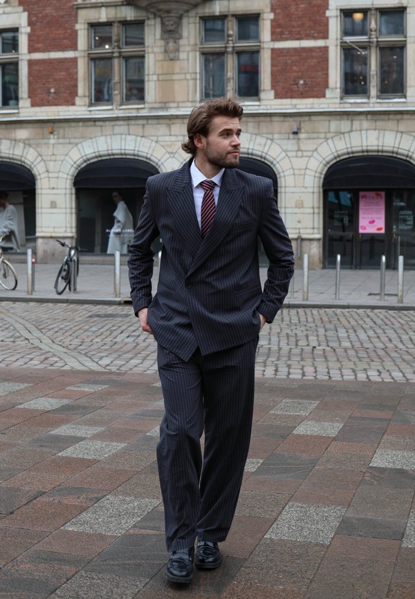 Mörk rutig kostym med slagkrage, parat med en vit skjorta och bordeauxfärgad slips. Svarta dressskor fulländar outfiten.
