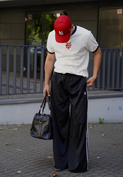 Camiseta blanca con ribete negro, logo rojo; pantalones de chándal negros con rayas blancas; gorra roja; bolsa negra con herrajes plateados.