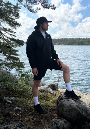 Chaqueta ligera negra con capucha sobre una camisa blanca, combinada con pantalones cortos negros, calcetines blancos y zapatillas negras. De pie sobre rocas junto al agua.