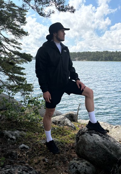 Chaqueta ligera negra con capucha sobre una camisa blanca, combinada con pantalones cortos negros, calcetines blancos y zapatillas negras. De pie sobre rocas junto al agua.