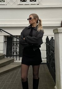 Γκρίζο κοντό μπουφάν με γιακά πάνω από μια μαύρη μπλούζα και μίνι φούστα. Διαφανείς μαύρες καλσόν και μαύρες μπότες μέχρι το γόνατο. Τα αξεσουάρ περιλαμβάνουν οβάλ γυαλιά ηλίου.